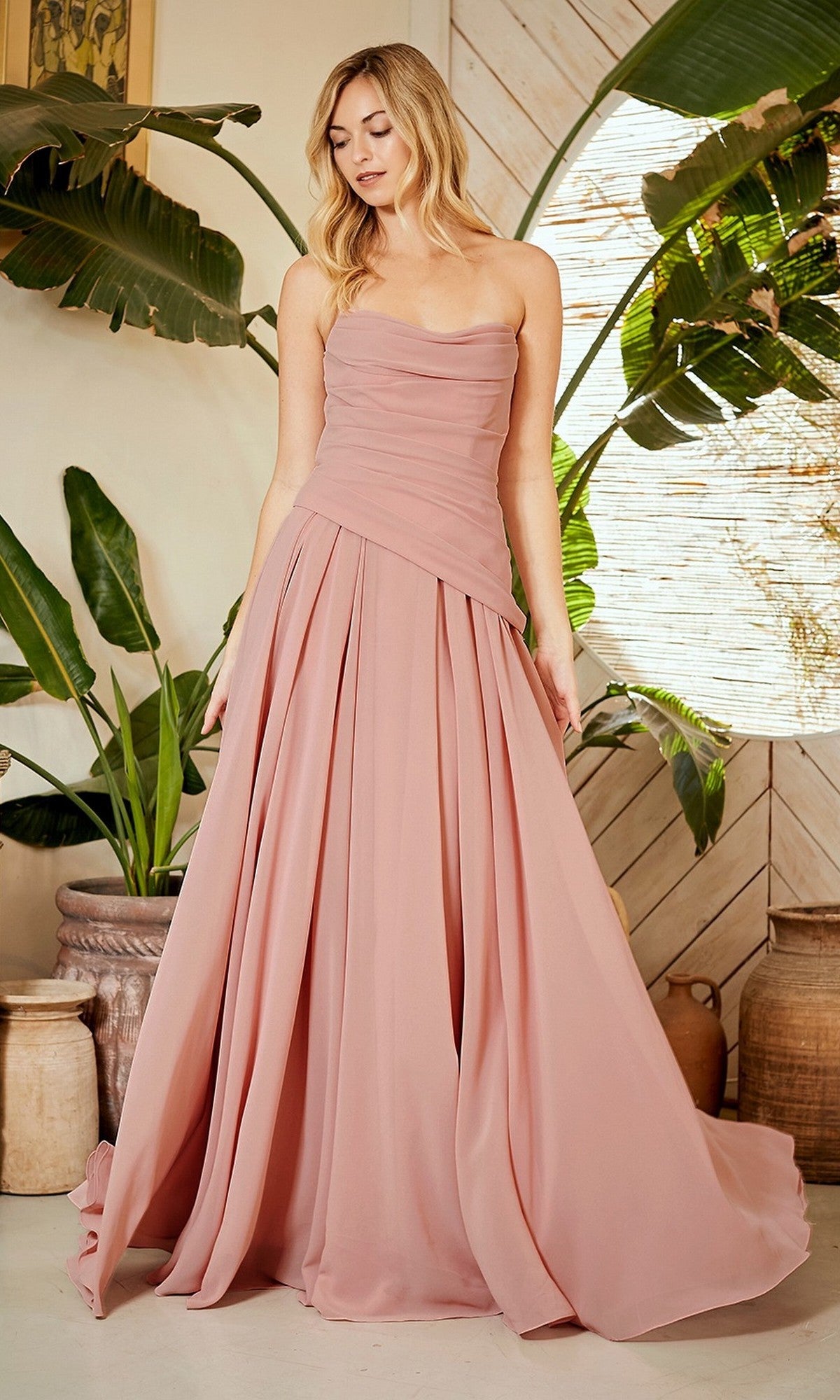 A woman stands indoors wearing the Amelia Couture 6133 long prom dress. The strapless blush pink chiffon gown features a pleated skirt and corset back. Green plants and large pottery are visible in the background.